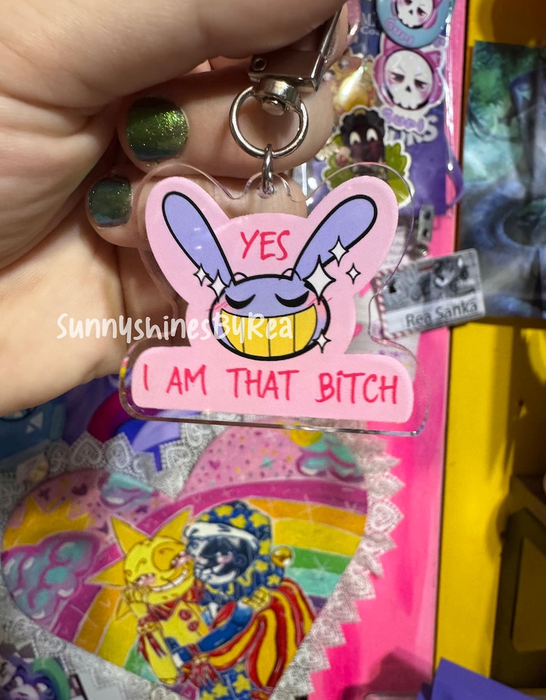 May include: A clear acrylic keychain featuring a cartoon character with purple ears and a yellow grin. The design includes the text YES and I AM THAT BITCH in pink and black lettering, accented with white sparkles on a light pink background.