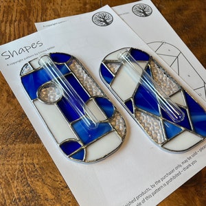 May include: Two stained glass bud vases with a geometric design in blue, white, and clear glass. Each vase has a clear glass tube for holding a single stem. The design is set against a white paper with the word "Shapes".