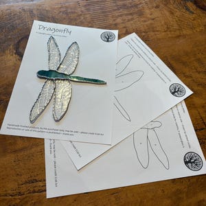 May include: A stained glass dragonfly pattern with instructions for creating a stained glass project. The pattern is on white paper with a green dragonfly on the top sheet. The dragonfly is outlined in black and has a green body. The instructions are on two sheets of paper with a black and white outline of the dragonfly.