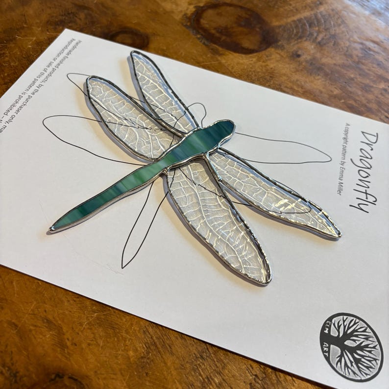 May include: A stained glass dragonfly with a green and white design. The dragonfly is on a white card with the text "Dragonfly" and "A copyright pattern by Emma Miller" printed on it.