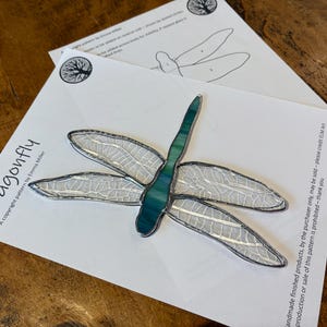 May include: A stained glass dragonfly pattern with a teal body and clear wings. The pattern is printed on white paper with the text "Dragonfly" and "A copyright pattern by Emma Miller".