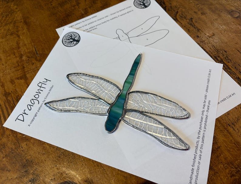 May include: A stained glass dragonfly pattern with a teal body and clear wings. The pattern is printed on white paper with the text "Dragonfly" and "A copyright pattern by Emma Miller".