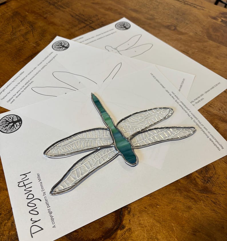 May include: A stained glass dragonfly pattern with a white and teal blue design. The pattern is printed on a white sheet of paper with the text "Dragonfly" and "A copyright pattern by Emma Miller".