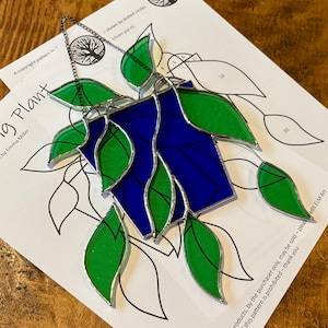 May include: A stained glass hanging plant with green leaves and a blue pot. The plant is hanging from a silver chain. The image includes a pattern for making the plant.