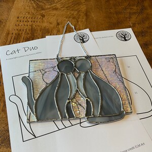 Peut inclure: Un duo de chats en vitrail, avec un cadre argenté, sur fond blanc. Les chats sont gris et blancs, et se font face. L'image comprend le texte "Cat Duo" et "A copyright pattern by Emma Miller".