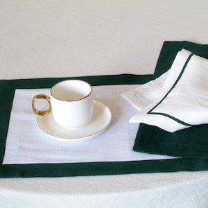 Peut inclure: Un set de table en lin blanc avec une bordure vert foncé et une serviette en lin blanc avec une bordure vert foncé. Une tasse et une soucoupe en porcelaine blanche sont sur le set de table.