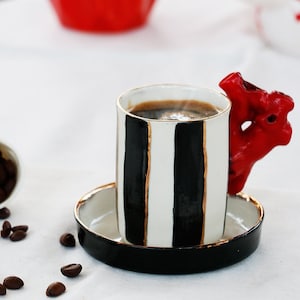 May include: A black and white striped coffee cup with a red, sculpted handle. The cup is filled with coffee and is sitting on a black saucer.