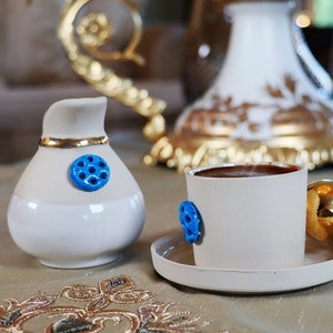 Könnte beinhalten: Eine weiße Kaffeetasse aus Keramik mit einem blauen Dekor und einem goldenen Henkel. Die Tasse steht auf einem weißen Unterteller. Die Tasse ist mit Kaffee gefüllt.