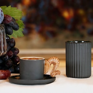 Könnte beinhalten: Eine schwarze Kaffeetasse mit einem braunen Henkel in Form eines Widderhorns. Die Tasse steht auf einem schwarzen Unterteller. Neben der Kaffeetasse steht eine schwarze Tasse mit geripptem Design.
