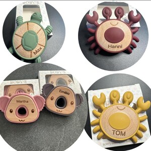 May include: A collection of baby teethers in various animal shapes. The teethers include a turtle, two crabs, and a koala, each with a wooden center and colorful silicone accents. Each teether has a name engraved on it.