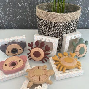 May include: A collection of baby teethers in the shapes of a koala, crab, and turtle. The teethers are made of wood and silicone in various colors, including brown, pink, and yellow. Each teether is packaged in a small box with the brand name "BOBY'S".
