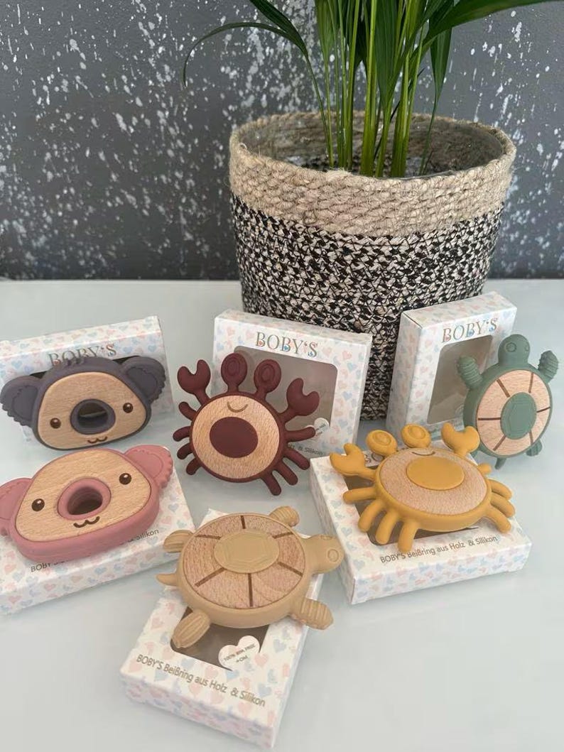 May include: A collection of baby teethers in the shapes of a koala, crab, and turtle. The teethers are made of wood and silicone in various colors, including brown, pink, and yellow. Each teether is packaged in a small box with the brand name "BOBY'S".