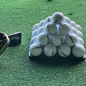 May include: A pyramid of white golf balls with black stripes on a black tray. The balls are arranged in a triangular shape with the top ball being the highest point. The tray is sitting on a green artificial grass surface. A black and silver golf club is in the lower left corner of the image.