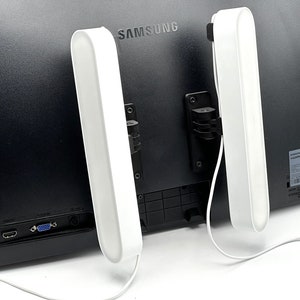 Op de afbeelding: Twee witte LED-lichtbalken bevestigd aan de achterkant van een zwarte computermonitor. De lichtbalken zijn met witte snoeren aan de monitor aangesloten.