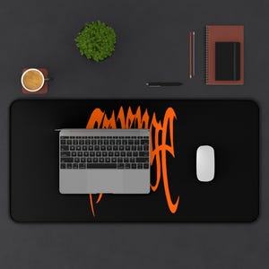 May include: Black desk mat with an orange graphic design and a laptop, mouse, and other office supplies. The mat features a stylized orange text design. A small plant, coffee cup, pen, and notebook are also visible.