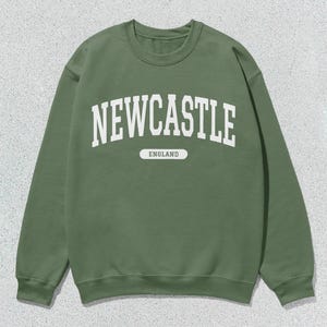 K&ouml;nnte beinhalten: Ein gr&uuml;ner Pullover mit wei&szlig;em Text, der "NEWCASTLE ENGLAND" lautet.