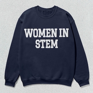 Peut inclure: Un sweat-shirt bleu marine avec le texte "WOMEN IN STEM" en lettres blanches en gras. Le sweat-shirt à col rond est présenté sur une surface texturée gris clair. Le haut à manches longues est un vêtement décontracté.
