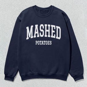 Puede incluir: Una sudadera azul marino con texto blanco que dice "MASHED POTATOES" en una fuente arqueada.