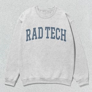 Może przedstawiać: Jasnoszara bluza z napisem "RAD TECH" w niebieskich literach na piersi. Bluza ma okrągły dekolt i długie rękawy. Tło to jasnoszara, teksturowana powierzchnia.