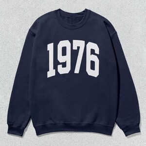 May include: Navy blue sweatshirt featuring the year "1976" in large, white, block lettering across the chest. The crew neck sweatshirt is a solid colour and has long sleeves.