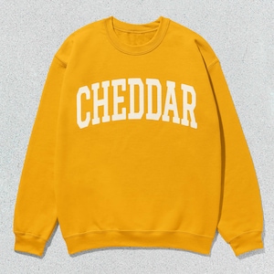 Puede incluir: Sudadera de color amarillo mostaza con la palabra "CHEDDAR" en letras blancas arqueadas. La parte superior de manga larga con cuello redondo está hecha de un material suave y es adecuada para ropa informal. La sudadera se muestra sobre un fondo gris claro.