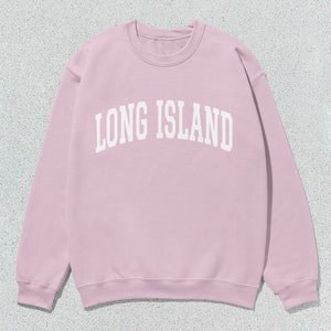 Pu&ograve; includere: Una felpa rosa chiaro con la scritta bianca "LONG ISLAND" in caratteri arcuati. La felpa girocollo &egrave; di colore uniforme e ha maniche lunghe. L'immagine &egrave; ripresa dall'alto.