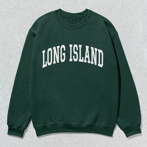 Pu&ograve; includere: Una felpa verde scuro con la scritta bianca "LONG ISLAND" ad arco. La felpa ha un girocollo e maniche lunghe. L'indumento &egrave; esposto su uno sfondo grigio maculato.