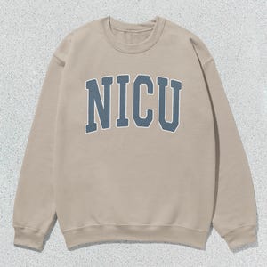 Puede incluir: Sudadera beige de cuello redondo con la palabra "NICU" en letras azules en el pecho. La sudadera tiene mangas largas y cuello acanalado. El fondo es una superficie moteada de color gris claro.