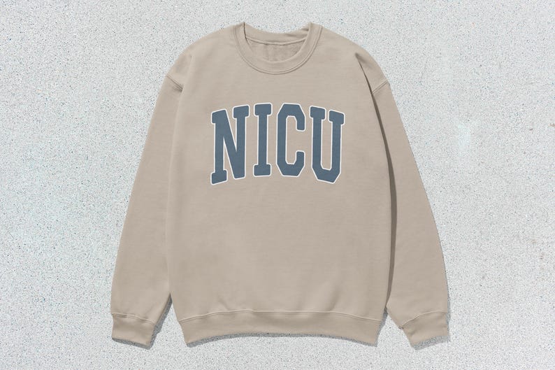 Puede incluir: Sudadera beige de cuello redondo con la palabra "NICU" en letras azules en el pecho. La sudadera tiene mangas largas y cuello acanalado. El fondo es una superficie moteada de color gris claro.