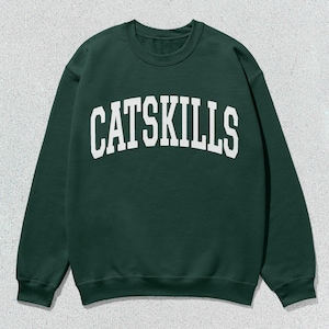 Könnte beinhalten: Dunkelgrünes Sweatshirt mit dem weißen Schriftzug "CATSKILLS" in großen, gebogenen Buchstaben auf der Brust. Das Sweatshirt hat einen Rundhalsausschnitt und lange Ärmel. Der Hintergrund ist hellgrau.