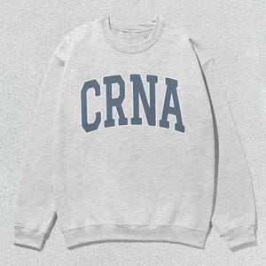 May include: A light gray sweatshirt with the word "CRNA" in blue block letters across the chest. The sweatshirt has a crew neck and long sleeves. The background is a light gray textured surface.