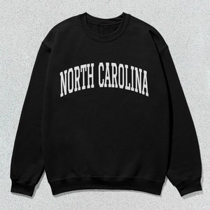 Pu&ograve; includere: Felpa nera con testo bianco che recita "NORTH CAROLINA" in un font ad arco.