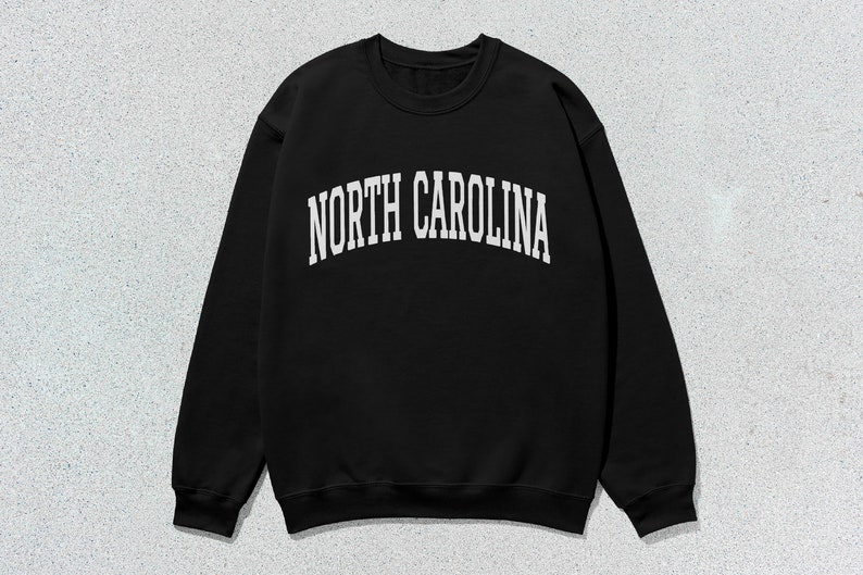 Pu&ograve; includere: Felpa nera con testo bianco che recita "NORTH CAROLINA" in un font ad arco.