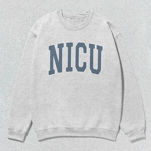 Puede incluir: Una sudadera gris claro con la palabra "NICU" en letras azules en el pecho. La sudadera de cuello redondo est&aacute; hecha de un material suave y tiene mangas largas. El fondo es una superficie texturizada gris claro.