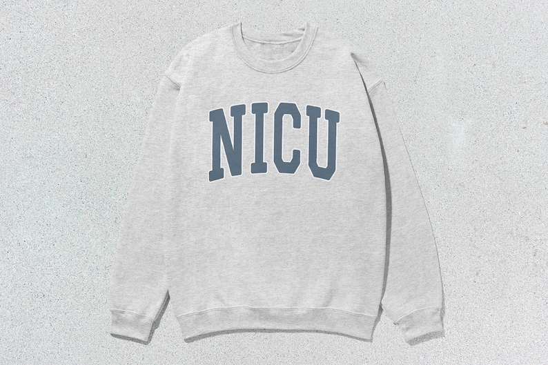 Puede incluir: Una sudadera gris claro con la palabra "NICU" en letras azules en el pecho. La sudadera de cuello redondo est&aacute; hecha de un material suave y tiene mangas largas. El fondo es una superficie texturizada gris claro.