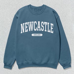 K&ouml;nnte beinhalten: Ein blauer Pullover mit wei&szlig;em Text, der auf der Vorderseite "NEWCASTLE ENGLAND" lautet.