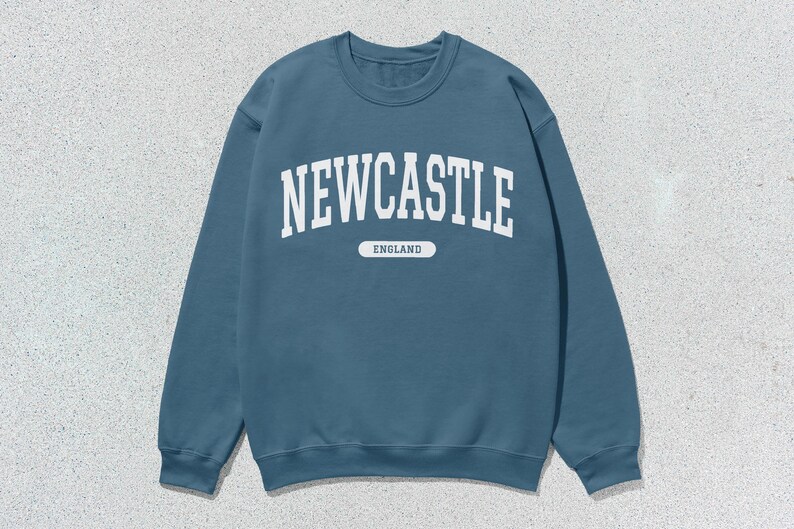K&ouml;nnte beinhalten: Ein blauer Pullover mit wei&szlig;em Text, der auf der Vorderseite "NEWCASTLE ENGLAND" lautet.