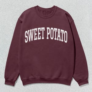 May include: A maroon sweatshirt with the text "SWEET POTATO" in white, arched lettering. The crew neck sweatshirt is made of a soft material and is suitable for casual wear.