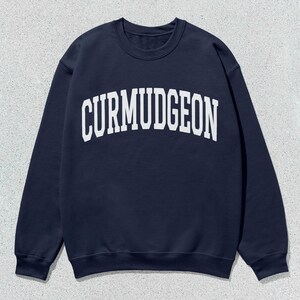 May include: Navy blue sweatshirt with the word "CURMUDGEON" printed in white, arched across the chest. The sweatshirt has a crew neck and long sleeves. The background is a light gray, textured surface.