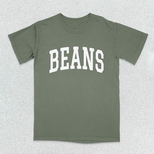 May include: A sage green short-sleeve t-shirt featuring the word BEANS printed in a large, white, arched, collegiate-style font across the chest. The garment has a crew neckline and a standard fit, showcasing a simple and clean design.