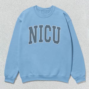 Puede incluir: Sudadera azul claro con la palabra "NICU" en gris, con una fuente de estilo universitario en el pecho. La sudadera tiene cuello redondo y mangas largas. El fondo es una superficie texturizada de color gris claro.