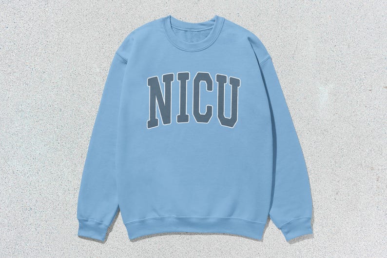 Puede incluir: Sudadera azul claro con la palabra "NICU" en gris, con una fuente de estilo universitario en el pecho. La sudadera tiene cuello redondo y mangas largas. El fondo es una superficie texturizada de color gris claro.