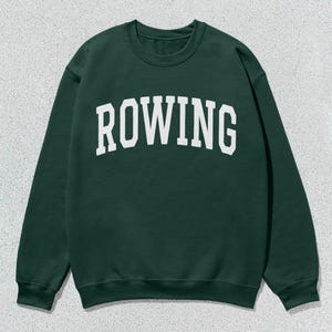 Puede incluir: Sudadera verde oscuro con la palabra "ROWING" impresa en letras blancas grandes y arqueadas en el pecho. La sudadera tiene cuello redondo y mangas largas, y se muestra sobre un fondo texturizado gris claro.