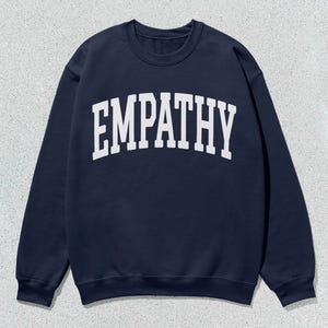 Può includere: Felpa blu navy con la scritta "EMPATHY" stampata a grandi lettere bianche arcuate sul petto. La felpa ha un girocollo e maniche lunghe. L'indumento è presentato su una superficie strutturata grigio chiaro.