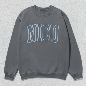 Puede incluir: Una sudadera gris con cuello redondo con la palabra "NICU" en una fuente azul y blanca de estilo universitario en el pecho. La sudadera tiene mangas largas y un cuello y pu&ntilde;os acanalados. El fondo es una superficie texturizada gris claro.