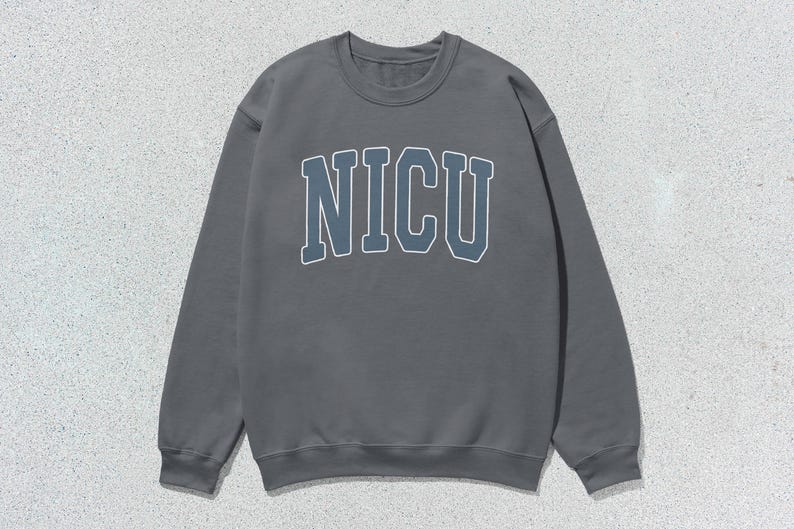 Puede incluir: Una sudadera gris con cuello redondo con la palabra "NICU" en una fuente azul y blanca de estilo universitario en el pecho. La sudadera tiene mangas largas y un cuello y pu&ntilde;os acanalados. El fondo es una superficie texturizada gris claro.