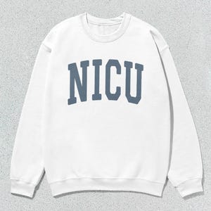 Puede incluir: Sudadera blanca de cuello redondo con la palabra "NICU" impresa en letras azules en el pecho. La sudadera tiene mangas largas y un cuello acanalado.