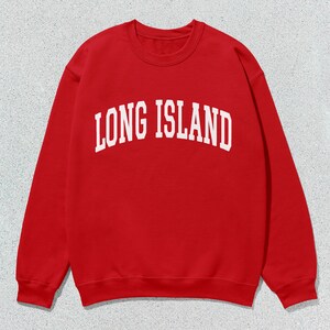 Pu&ograve; includere: Una felpa rossa con la scritta bianca "LONG ISLAND" in caratteri arcuati. La felpa ha un girocollo e maniche lunghe. L'immagine &egrave; ambientata su uno sfondo grigio maculato.
