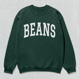 May include: A dark green sweatshirt featuring the word "BEANS" in large, white, block letters. The sweatshirt has a crew neck and long sleeves. The background is a light grey, textured surface.