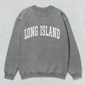 Pu&ograve; includere: Una felpa grigia con la scritta "LONG ISLAND" in bianco, a lettere arcuate. La felpa girocollo &egrave; realizzata in un materiale morbido ed &egrave; adatta per un abbigliamento casual. L'immagine &egrave; scattata su uno sfondo strutturato grigio chiaro.